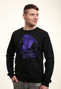 Man in een zwart sweatshirt met een paarse afbeelding van een meisje met vlechten en de tekst "Sub-Urban Gothic," staand met één hand in zijn zak.