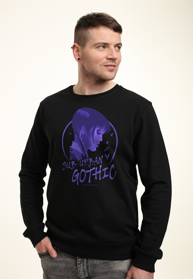Man in een zwart sweatshirt met een paarse afbeelding van een meisje met vlechten en de tekst "Sub-Urban Gothic," staand met één hand in zijn zak.