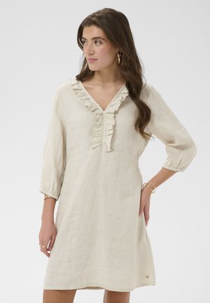 Kvinde med langt brunt hår iført en beige knælængde kjole med flæset V-udskæring og trekvart ærmer, poserede med den ene hånd på hoften.