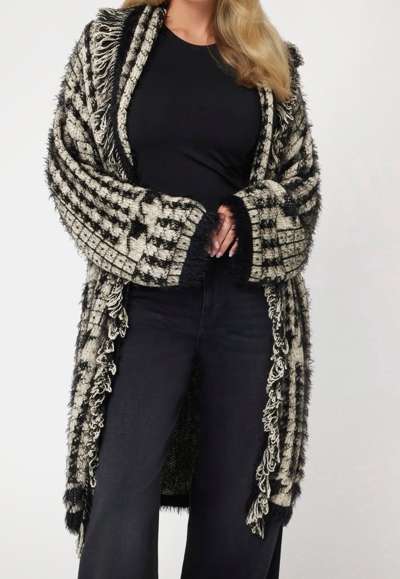 Donna che indossa un top e pantaloni neri, con un lungo cardigan a frange checkered nero e beige avvolto attorno alle spalle.