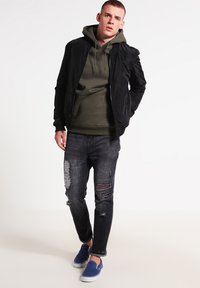 Zwarte bomberjack over een olijfgroene hoodie, gecombineerd met gescheurde grijze jeans en blauwe instap schoenen. Casual, moderne ontwerpelementen.