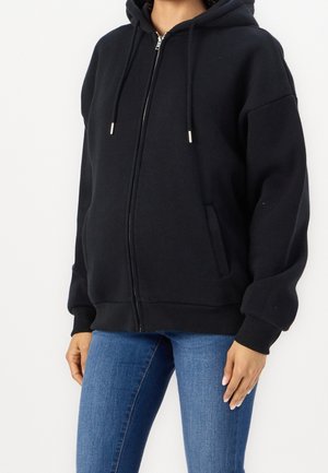 Personne portant un sweat à capuche noir zippé avec des cordons et un jean bleu, montrée du cou aux hanches sur un fond uni.