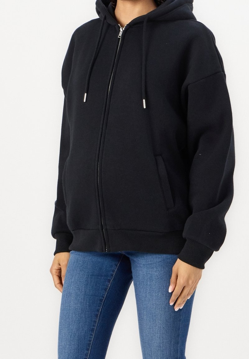 Personne portant un sweat à capuche noir zippé avec des cordons et un jean bleu, montrée du cou aux hanches sur un fond uni.