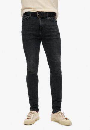 Zwarte slim-fit jeans met subtiele vervaging, vijf-pocket ontwerp en een zwarte riem met zilveren gesp. Gecombineerd met beige sneakers.