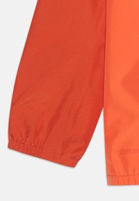 Manica di giacca impermeabile arancione con polsini elastici, texture liscia e una leggera personalizzazione sulla stoffa. Design compatto e moderno.