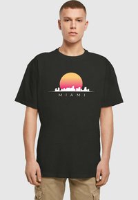 Svart bomull t-skjorte med et grafisk trykk av en solnedgang og silhuett av byens skyline med ordet "MIAMI" i hvitt. Korte ermer, avslappet passform.