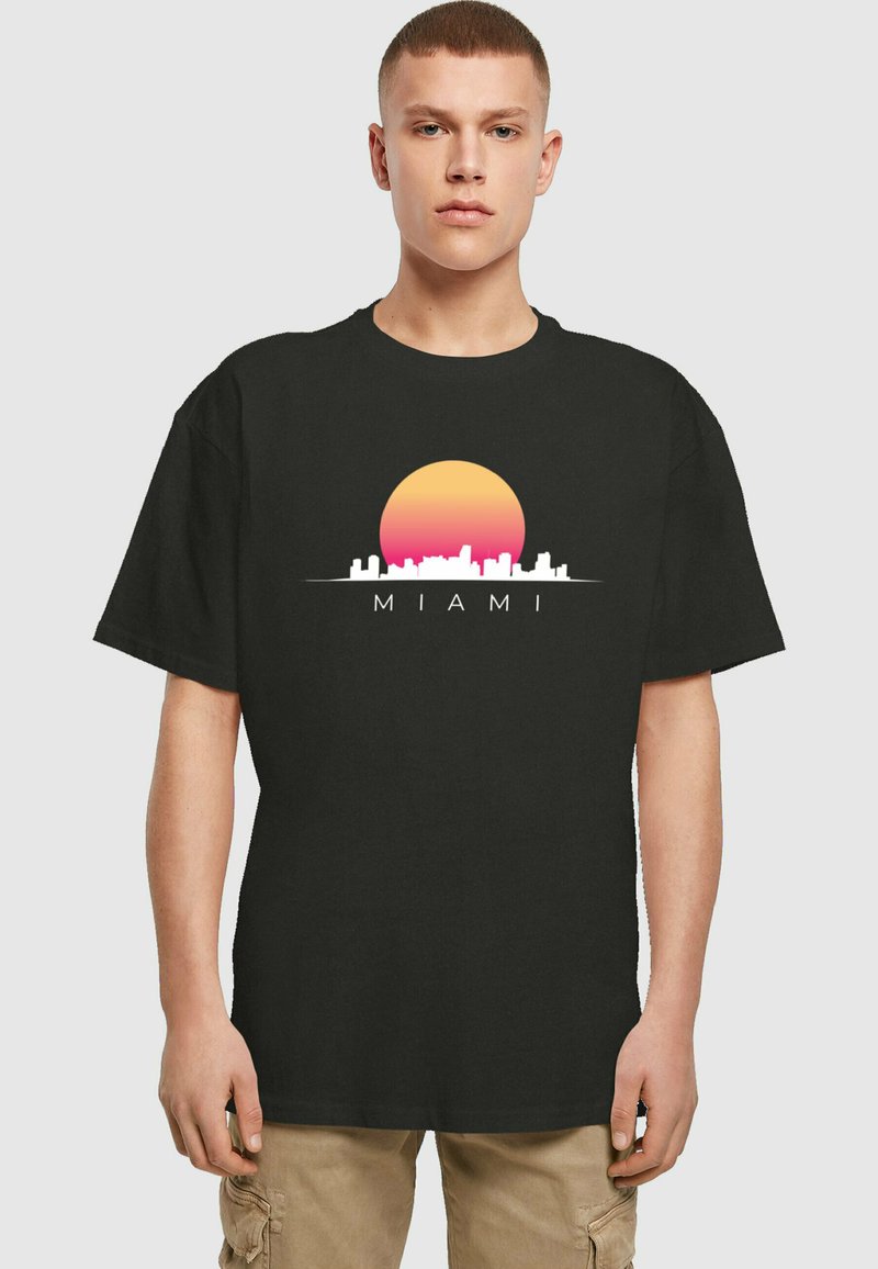 Svart bomull t-skjorte med et grafisk trykk av en solnedgang og silhuett av byens skyline med ordet "MIAMI" i hvitt. Korte ermer, avslappet passform.
