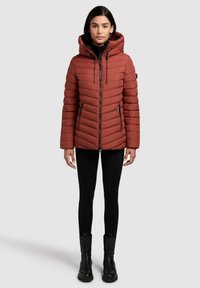 Rust-colored puffer jacket with a hood, featuring vertical quilting, front zipper, and two side pockets. Worn with black leggings and ankle boots.