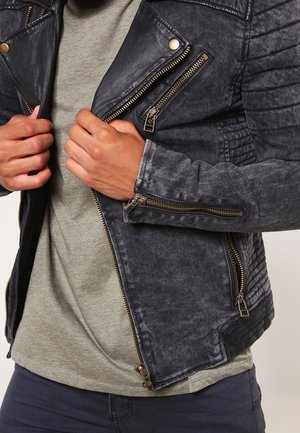 Mains ajustant la fermeture éclair avant d'une veste en cuir noire texturée portée par-dessus une chemise gris clair et un pantalon foncé.