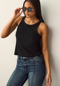 Schwarzes, geripptes Tanktop kombiniert mit hochtaillierten blauen Jeans. Das Top hat einen runden Ausschnitt und großzügige Armausschnitte. Das Model trägt dunkle Sonnenbrillen.