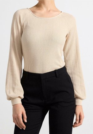 Vrouw draagt een beige ribgebreide trui met ballonmouwen en zwarte hoge taille broek, staand tegen een witte achtergrond.