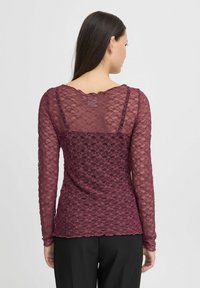 Bordeaux-rotes langärmliges Spitzenshirt mit scallopförmigem Ausschnitt, transparenter Textur und floralem Muster. Getragen mit schwarzen Hosen.
