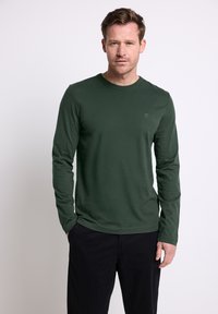 Długosleeve'owa koszula w ciemnozielonym kolorze, wykonana z miękkiego materiału. Posiada okrągły dekolt i małe logo na piersi. Połączona z czarnymi spodniami.