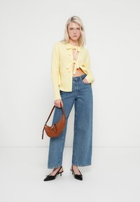 Kollane kootud kardigan lipsudetailiga, kombineeritud kõrge vöökohaga siniste laiade jalgadega teksade ning mustade teravate kontsadega. Pruun käekott.