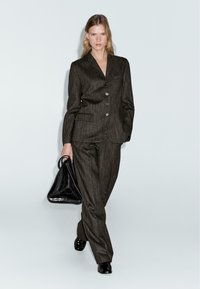 Grijze streepjesstof pak met een op maat gemaakte blazer en losse broeken. Het model draagt een zwarte driehoekige handtas en heeft glanzende zwarte schoenen aan.