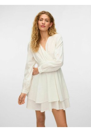 Donna con capelli biondi ricci che indossa un vestito bianco a portafoglio con maniche lunghe e gonna a strati, che posa su uno sfondo semplice.