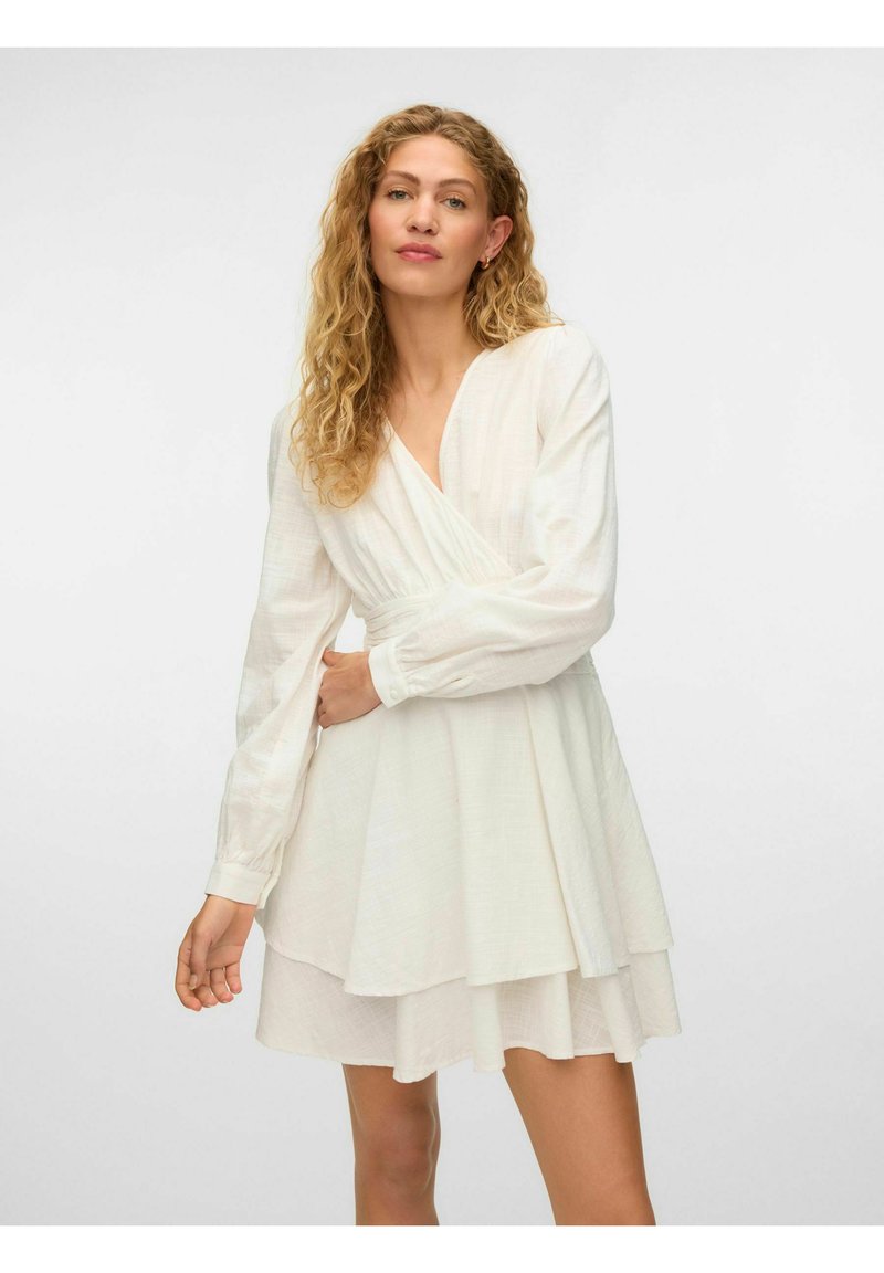Femeie cu păr blond creț purtând o rochie albă tip wrap cu mâneci lungi și fustă stratificată, pozând pe un fundal simplu.