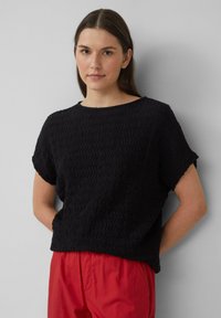 Haut noir à manches courtes, texturé avec un motif ondulé. Encolure ras du cou, coupe décontractée. Assorti avec un pantalon rouge. Fond neutre.