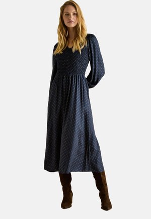 Marineblaue langärmlige Kleid mit gerafftem Oberteil, gepunktetem Muster und fließendem Rock. Kombiniert mit kniehohen braunen Wildlederstiefeln.