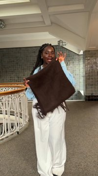 Rjava, resasta kvadratna ruta, ki jo drži v roki, narejena iz mehkega materiala. Model nosi svetlo modro pulover in bele široke hlače v sodobnem notranjem prostoru.