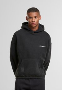 Giovane in felpa nera con la scritta "Sunday Running Club", in piedi con le mani dietro la schiena su uno sfondo grigio uniforme.