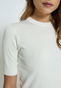 Ljust beige ribbat stickad topp med rund halsringning och korta ärmar med texturerade manschetter. Slät tyg med en kroppsnära passform.