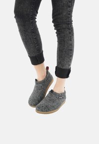 Graue Filzschlappen mit runder Form, Gummisohlen und dekorativen Knöpfen, getragen mit dunkelblauen Jeans, die an den Bündchen hochgekrempelt sind.