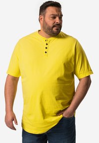 Camisa henley amarilla de manga corta con un placket de dos botones y un ajuste relajado, combinada con jeans oscuros, presentada sobre un fondo blanco.