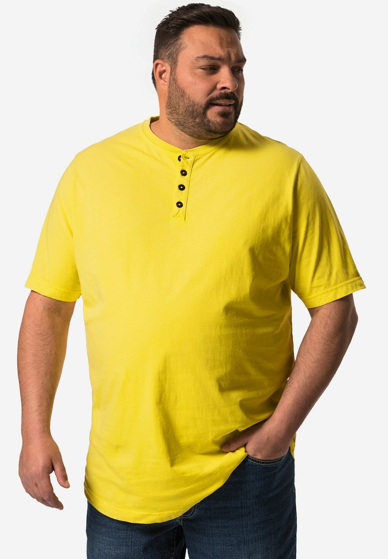Camisa henley amarilla de manga corta con un placket de dos botones y un ajuste relajado, combinada con jeans oscuros, presentada sobre un fondo blanco.