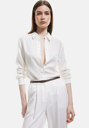Camicia bianca con colletto, tessuto strutturato e scollo profondo. Abbinata a pantaloni bianchi e una cintura di pelle marrone.