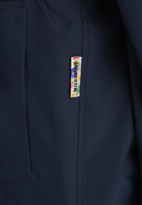 Marineblauber Stoff mit glatter Textur, verfügt über ein dekoratives Etikett mit floralen Mustern und "MILES ONE" in Schwarz gedruckt.