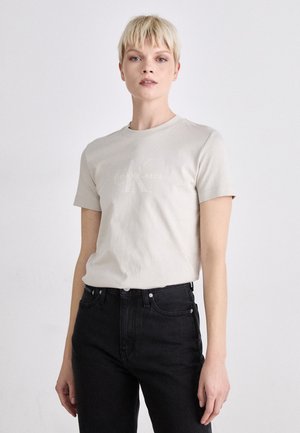 Šviesiai smėlio spalvos medvilninė marškinėliai trumpomis rankovėmis, su iškilniu "Calvin Klein Jeans" logotipu priekyje. Sujungti su juodais džinsais.