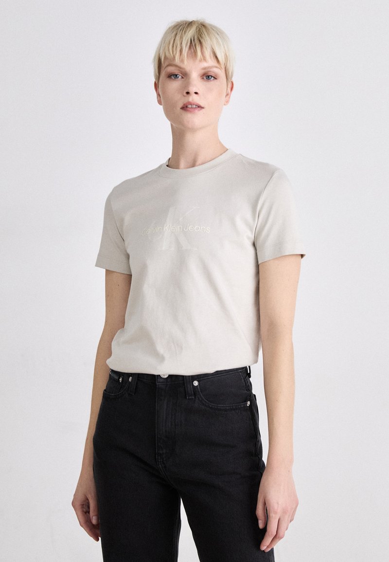 Svetlo béžové tričko z bavlny s krátkymi rukávmi, s vyrazeným logom "Calvin Klein Jeans" na prednej strane. V kombinácii s čiernymi džínsami.