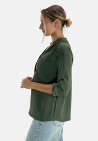 Blusa verde scuro a tre quarti in tessuto leggero, con un bottone a tab, scollatura a V e taschino sul petto, abbinata a jeans chiari.