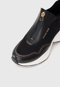 Sorte og brune sneakers med åndbart mesh-overdel, glidelås-lukking og hvit midtsåle med teksturdetaljer.