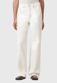 Jeans bianchi a gamba larga realizzati in denim, con tasche anteriori, chiusura a bottone e cerniera, e una consistenza liscia. Sono visibili anche scarpe con borchie.