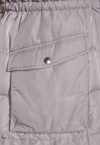 Ljusgrå pufferjacka i tyg med en stor, knäppt knappficka och synlig sömnad. Ytan är slät med en subtil glans.