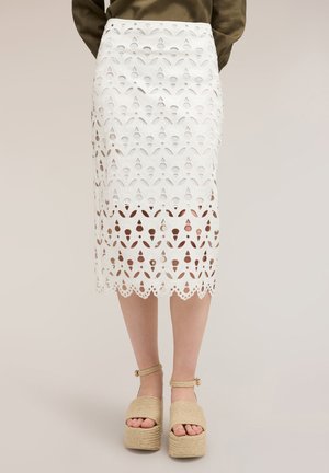 Hvid knælang nederdel med blomsterudskåret mønster båret med beige platformsandaler og en grøn top, vist fra talje til fødder.