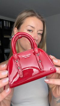 Κόκκινη τσάντα από δερμάτινο βερνίκι με δομημένο σχήμα, διαθέτει διπλές λαβές, κλείσιμο με φερμουάρ και ασημένια πινακίδα με το λογότυπο.