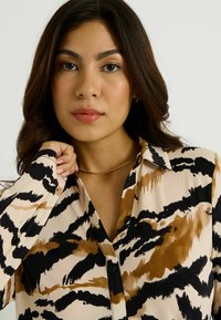 Camisa de botón con base beige y estampado abstracto de animales en negro y marrón. Mangas largas y cuello estándar. Textura de tela suave.