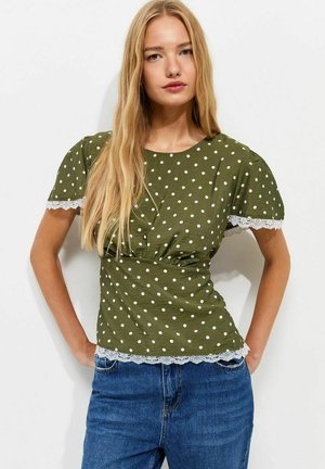 Jonge vrouw met lang blond haar, die een olijfgroene polkadot blouse met kanten rand en een blauwe spijkerbroek draagt, staand met handen in de zij.