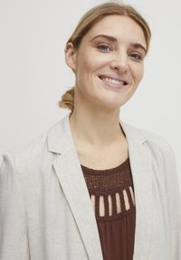 Harmaa beige-bleiseri, jossa on teksturoitu pinta, ruskean yläosan päällä, jossa on virkattuja yksityiskohtia ja aukkoja. Sileä kangas ja muotoiltu malli.