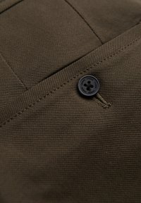 Tessuto di cotone marrone con una tessitura diagonale, caratterizzato da un dettaglio con bottone nero cucito su una tasca. La trama sembra resistente e durevole.