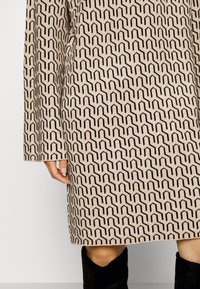 Abito beige con motivo, realizzato in tessuto texturizzato, caratterizzato da un design geometrico nero. Maniche lunghe con vestibilità rilassata e dettaglio con spacco laterale.