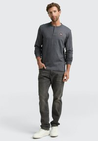 Grau langärmliges Henley-Shirt mit Knopfausschnitt und rundem Logo, kombiniert mit dunklen Jeans und weißen Turnschuhen vor einem hellen Hintergrund.