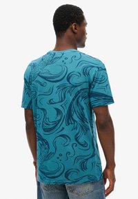 Camiseta de algodón turquesa con un diseño de remolino azul y patrón de peces. Manga corta, cuello redondo y un corte relajado.
