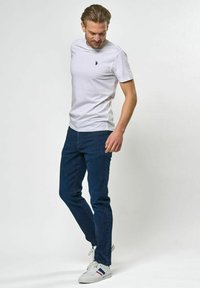 Balta kokvilnas T-krekls ar mazu tumši zilu logotipu uz krūtīm, kombinēts ar tumši zilām džinsa biksēm un baltām sporta kurpēm ar izceltiem svītrām.