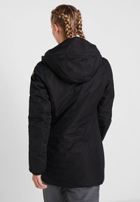 Osoba s copánky, oblečená v černé mikině s kapucí, zobrazená zezadu na světlejším jednobarevném pozadí.