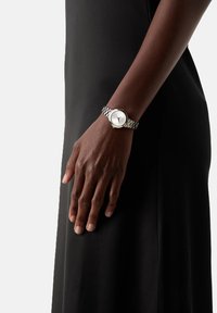 Sidabrinė apyrankė su apvaliu baltu ciferblatu, minimalistiškais skaičiais ir metaline grandinine apyranke, pavaizduota ant rankos prieš juodą foną.