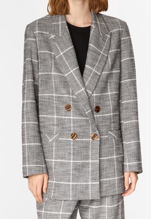 Femme portant un blazer croisé à carreaux gris avec des boutons marron sur un top noir et un pantalon à carreaux assorti.